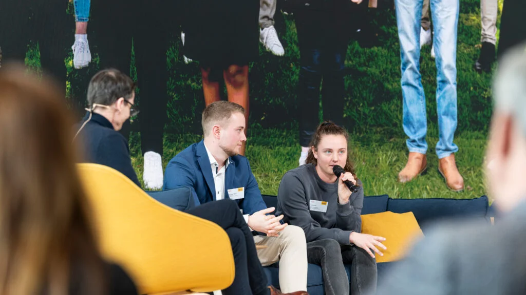 Christoph Mayer von Dachser und Lara Kühn von ZAK sitzen auf einem Sofa auf der Bühne und Lara spricht ins Mikrofon