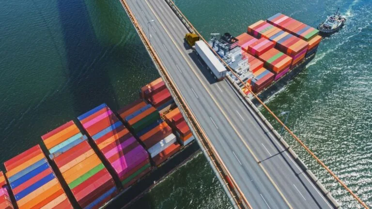 Weiß-gelber Lkw fährt auf einer Brücke, unter der ein Containerschiff fährt