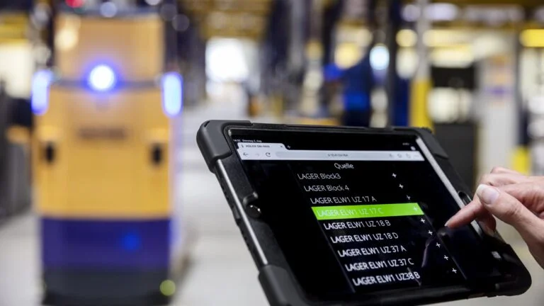 AGV_and_Tablet_2048_1152 Tablet mit Lagerverwaltungsanwendung im Vordergrund, während im Hintergrund ein fahrerloses Transportfahrzeug (AGV) in einer Lagerhalle unterwegs ist.