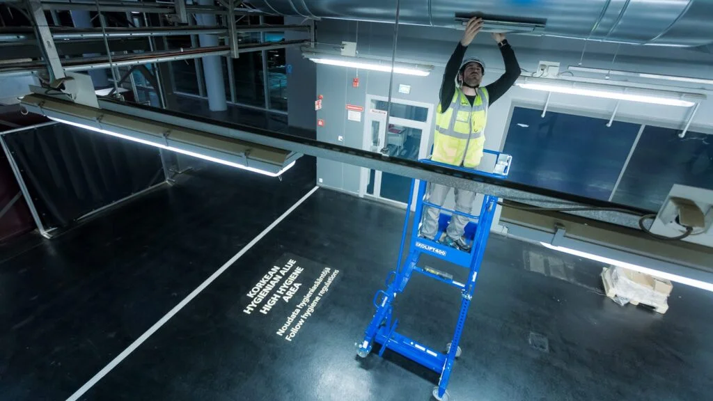 Ein Mann in Arbeitskleidung steht auf einer Ixolift Arbeitsbühne in einer Industriehalle und montiert Beleuchtung unter der Decke.